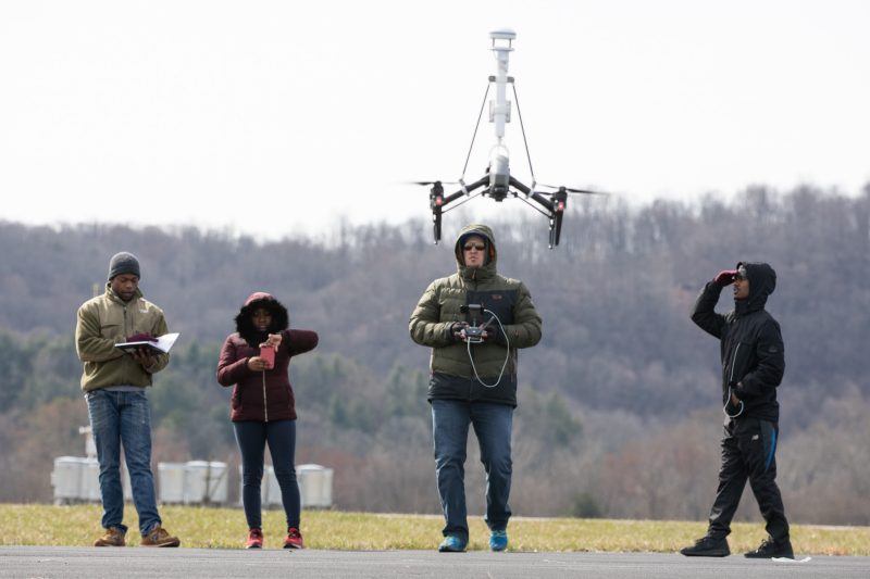 Undergraduates from Bennett, Morehouse, and Hampden Sydney stand on an airstrip at Virginia Tech’s Kentland Farm. David Schmale is manning a drone. One student is writing in a notebook, another is recording a video on her phone, and another is observing the drone and protecting his eyes with his hands. Courtesy of Peter Means.